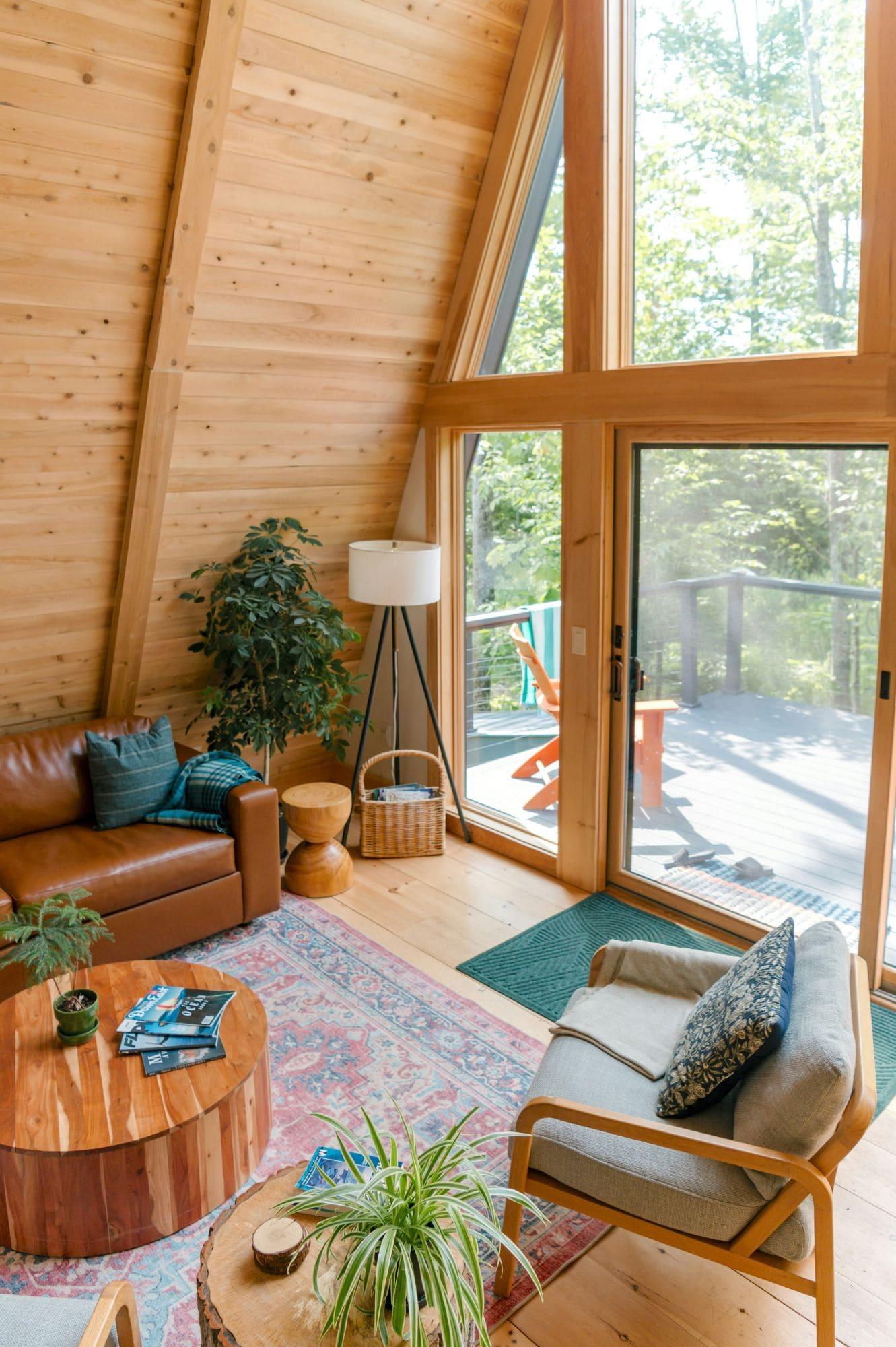 A sunlit A-frame living room in pale wood, with a leather sofa, a rug and tall windows onto a deck.
